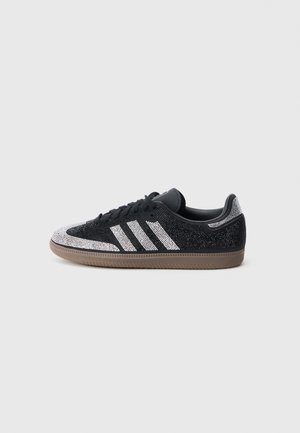 Czarny niski tenis z białymi paskami i ozdobami z kamieniami na brązowej podeszwie na jednolitym jasnoszarym tle.