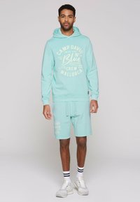 Conjunto de sudadera con capucha y pantalones cortos en color turquesa, mezcla de algodón, con texto y logo impresos en blanco. Ajuste casual con puños acanalados y cintura con cordón.