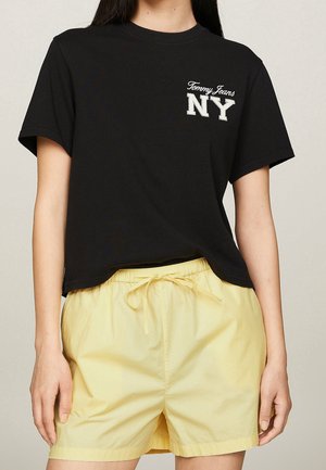 Frau trägt ein schwarzes Kurzarmshirt mit weißem Text ""Tommy Jeans NY"" und hellgelben Shorts mit Tunnelzug vor neutralem Hintergrund.