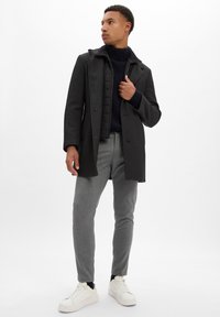 Manteau noir au design structuré, fermeture à boutons et poches plaquées, associé à un col roulé côtelé bleu marine et à un pantalon gris fuselé. Baskets blanches.
