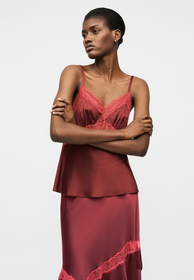 Vrouw met kort haar, gekleed in een bordeauxroze satijnen camisol met kanten rand en bijpassende rok, met gekruiste armen, kijkend naar de zijkant.
