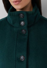 Grüne Fleecejacke mit hohem Kragen und vier schwarzen Knöpfen. Der Stoff hat eine weiche Textur und zeigt eine dezente, einfarbige Optik.