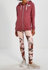 Maroon zip-up hoodie with white logo, paired with pink camo-patterned leggings featuring red and gray accents, and light-colored sneakers.