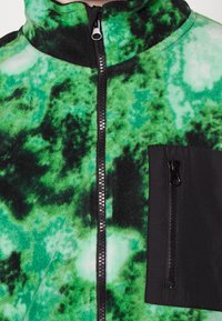 Giacca in pile con un motivo astratto verde e nero, colletto alto, zip frontale completa e tasca sul petto con zip in tessuto nero.