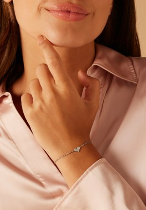 Femme portant un chemisier en satin rose clair touche son menton du doigt, montrant un bracelet chaîne argentée avec un pendentif en forme de cœur au poignet.