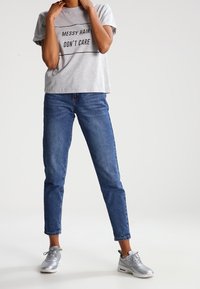 Camiseta de algodón gris con el texto negro "EL CABELLO DESORDENADO NO ME IMPORTA", combinada con jeans azules y zapatillas plateadas, mostrando un atuendo casual.