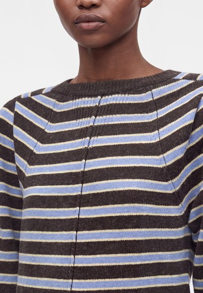 Femme portant un pull en tricot à rayures marron, bleu et beige avec un col rond. Seuls le bas du visage et le haut du torse sont visibles.