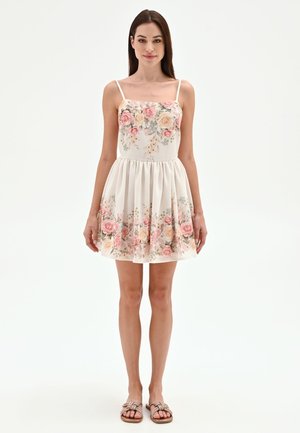 Femme debout portant une robe blanche sans manches avec un imprimé floral rose et beige et des sandales plates beiges sur fond blanc.