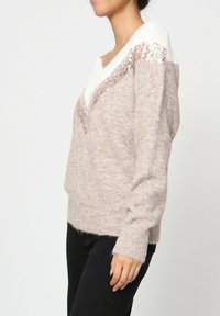 Un pull rose clair avec un profond décolleté en V, orné d'accents blancs et de sequins. Fabriqué en tissu tricoté doux et texturé avec des manches longues.