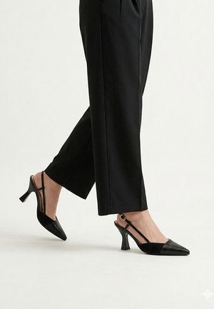 Persona que lleva zapatos de tacón slingback negros con punta afilada, tacones curvos de altura media y pantalones negros de pierna recta sobre un fondo claro.