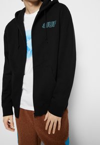 Persona con un hoodie nero con zip e logo blu "HUF", una maglietta grafica bianca e pantaloni in pile marroni con toppe blu.