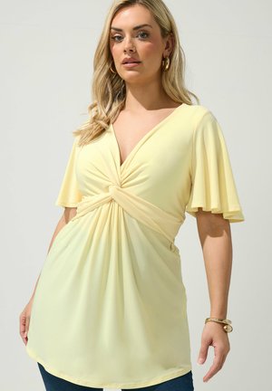 Frau mit langen blonden Haaren, die ein blassgelbes, kurzärmeliges Top mit gedrehtem Knoten sowie goldene Creolen und ein Armband vor einfarbigem Hintergrund trägt.