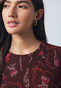 Pull marron avec des motifs graphiques festifs rouges et roses, incluant des boissons et des confettis, avec un col rond côtelé.