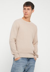Suéter de punto acanalado beige con cuello redondo, mangas largas y dobladillo ajustado, combinado con jeans de color azul claro.
