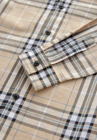 Chemise à carreaux beige en coton avec des rayures bleues, noires et blanches. Elle présente des manches longues, des poignets à boutons et des boutons foncés.