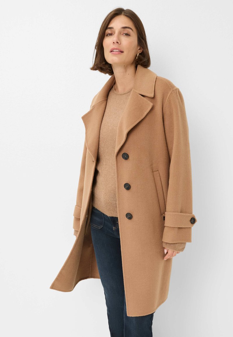 Manteau en mélange de laine camel avec de grands revers, fermeture à double boutonnage et poches latérales. Associé à un pull beige et un jean noir.