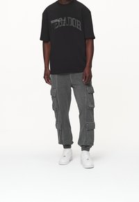 Mies, jolla on päällään musta ylisuuri t-paita, jossa on teksti "PEGADOR", harmaat cargo-housut taskuilla ja valkoiset lenkkarit, seisoo tasaisella valkoisella taustalla.