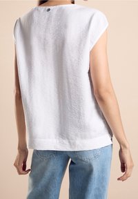 Top blanco de manga corta con acabado texturizado, escote redondeado y un pequeño detalle de logo en la parte trasera, combinado con jeans azul claro.