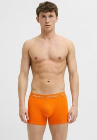 Jonge man die recht vooruit staat, shirtloos, met feloranje boxershorts met een "JACK & JONES" tailleband tegen een effen achtergrond.