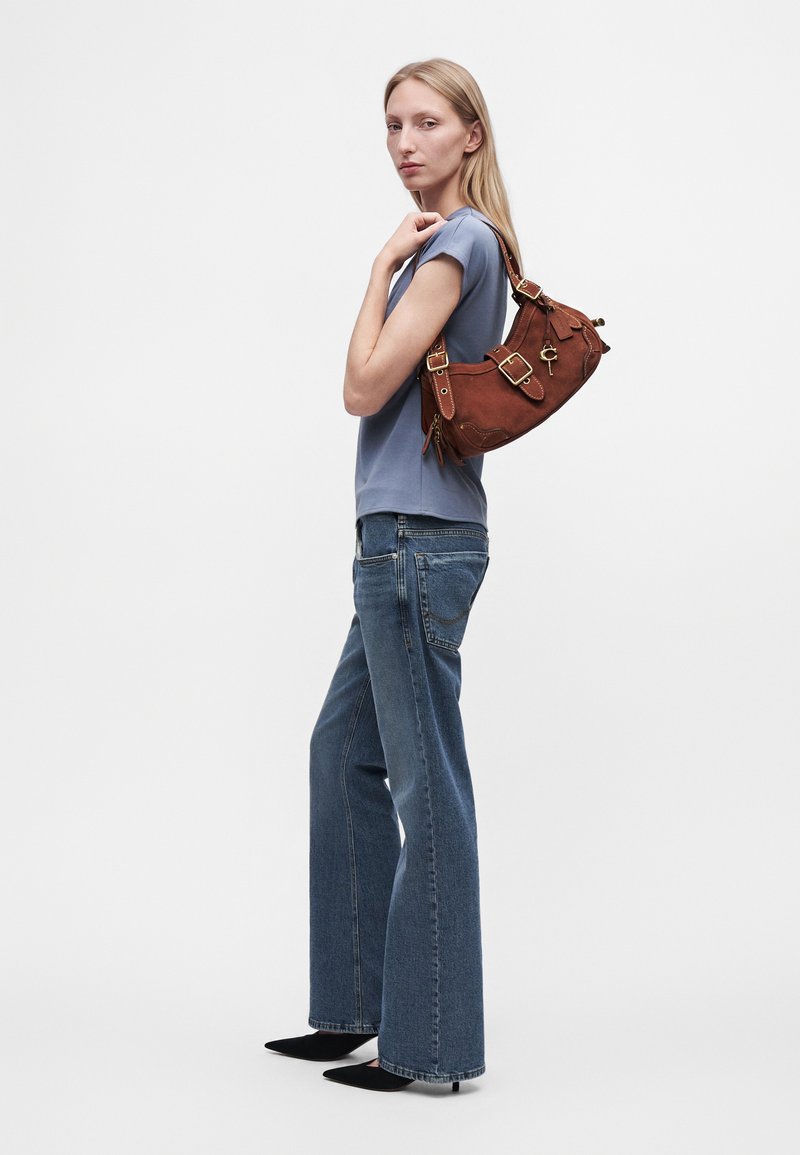 Femme aux longs cheveux blonds portant une chemise bleue, un jean à jambes larges et des talons noirs, portant un sac à bandoulière en cuir marron avec des boucles.