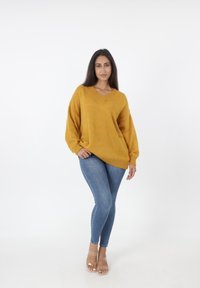 Mosterdgele oversized gebreide trui met een V-hals, lange mouwen en geribde zoom, gecombineerd met aangesloten blauwe jeans en doorzichtige sandalen met hakken.