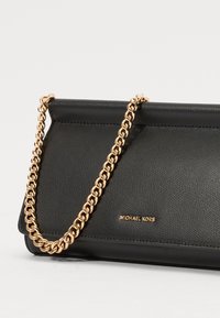Sac à main en cuir noir avec une bandoulière en chaîne dorée, présentant une texture lisse et un logo de marque en relief sur le devant.