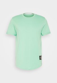 Tyrkysové triko s krátkým rukávem z měkkého materiálu, s kulatým výstřihem a štítkem s logem na levé straně blízko lemu.