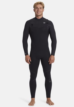 Young man standing barefoot, wearing a full black wetsuit with knee pads and a chest zipper against a plain white background.