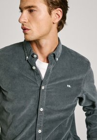 Grijze corduroy overhemd met knopen en een kraag, met een klein wit logo en witte knopen, gelaagd over een wit T-shirt.