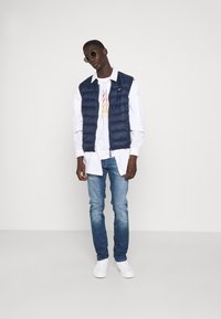 Chaleco acolchado azul marino sobre una camiseta de manga larga blanca con detalles impresos, combinado con jeans azul claro y zapatillas blancas, complementado con gafas de sol.