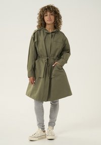 Jachetă lungă de culoare verde măsliniu, cu mâneci lungi și talie cu șnur, două buzunare frontale, o glugă și o textură netedă. Purtați cu blugi gri și adidași albi.
