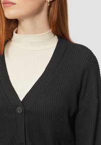 Schwarze gerippte Strickjacke mit V-Ausschnitt und Knopfverschluss, über einem weißen, figurbetonten Rollkragenpullover getragen. Glatte Textur und lockere Passform.