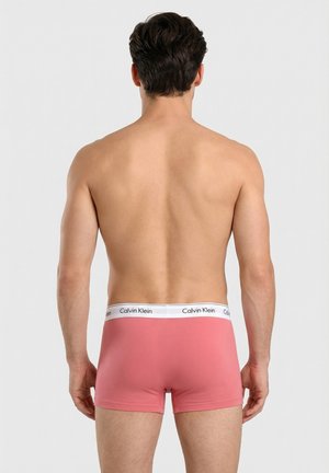 Hombre con cabello corto y oscuro, espalda desnuda, vistiendo calzoncillos bóxer rosas de Calvin Klein, de pie frente a un fondo claro y liso.