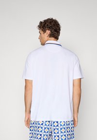 Hvit kortermet polo skjorte med klassisk krage, prydet med blå og gule striper langs kragen, og brukt sammen med mønstrede shorts.