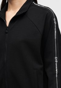 Μαύρο μπουφάν με φερμουάρ μπροστά, ψηλό γιακά και πλαϊνές τσέπες. Διαθέτει υφή υφάσματος, αντίθετες λευκές και γκρι ρίγες και λεπτομέρειες με λογότυπο.