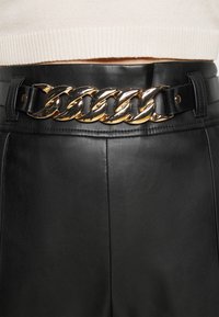 Svart läderbälte med en kraftig guldfärgad kedjespänne, slät yta och kontrasterande svarta metalldetaljer längs remmens kanter.