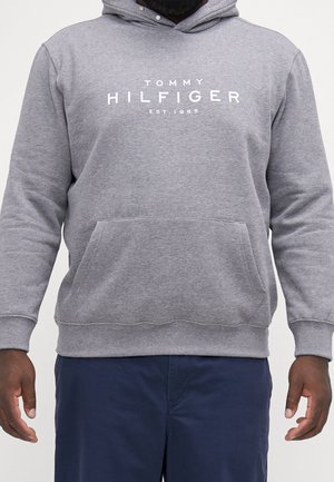 Mann trägt grauen Tommy Hilfiger Hoodie mit Fronttasche und marineblauer Hose, steht mit entspannten Armen neben dem Körper.