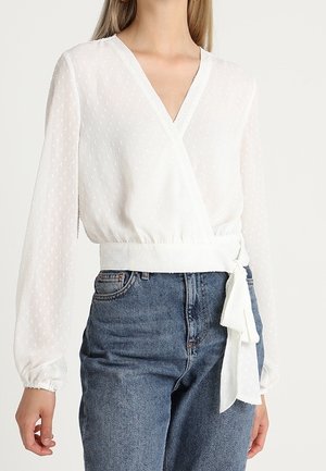 Blouse blanche à manches longues en tissu léger avec une texture subtilement ponctuée, col en V, poignets élastiques, ceinture latérale, portée avec un jean bleu taille haute.