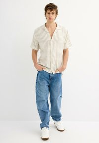 Camisa de manga corta a rayas en crema y azul, combinada con vaqueros azules de corte holgado y zapatillas blancas, de pie contra un fondo liso.