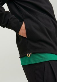 Črna sweatshirt s stranskimi žepi, ki vključuje zeleno majico spodaj. Ima oranžne detajle na robu s prilagoditvijo na vrvico.