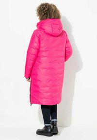 Abrigo largo de plumas rosa con capucha, que presenta un textura acolchada, cremalleras laterales y un corte holgado, combinado con jeans oscuros y botas negras de tobillo.