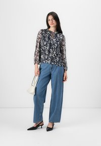 Blusa a fiori nei toni del blu e crema, con maniche lunghe in tessuto trasparente e scollatura a chiave. Abbinata a jeans blue a vita alta e tacchi neri.