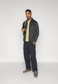 Ανδρικό σκούρο γκρι μπουφάν με φερμουάρ και υφή, ταμπά μπλούζα, μαύρο cargo παντελόνι και μαύρα παπούτσια πεζοπορίας με χρυσές λεπτομέρειες.