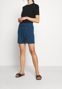 Modré kraťasy s šňůrkovým páskem a dvěma bočními kapsami, v kombinaci s černým topem s krátkým rukávem a černými pantofli s stříbrnou sponou.