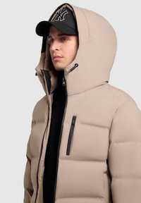 Beige steppelt kabát magas gallérral és állítható kapucnival. Fekete cipzárral és kiegészítőkkel rendelkezik, fekete felsőre és sapkára húzva.
