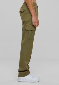 Olivengrüne Cargo-Hosen mit einem elastischen Bund, seitlichen Taschen und einer lockeren Passform, kombiniert mit weißen Sneakers. Die Textur wirkt glatt.