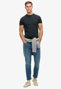 Sort t-shirt med korte ærmer, blå jeans, lys grå strikket sweater bundet om taljen, beige sneakers, stående på en ensfarvet hvid baggrund.