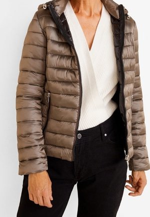 Femme portant une veste matelassée marron brillante par-dessus un pull blanc à col en V et un pantalon noir, debout avec les bras détendus le long du corps.