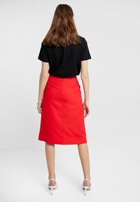 Falda lápiz roja ajustada con cintura alta y abertura trasera, combinada con una blusa de manga corta negra y sandalias de tacón blancas.
