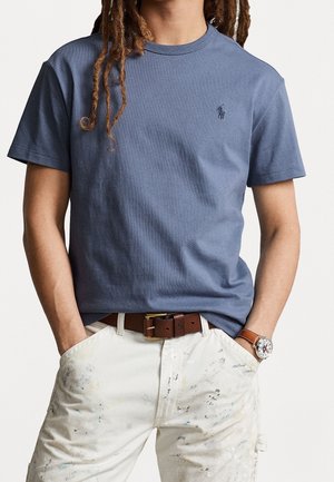 Persona che indossa una T-shirt blu a maniche corte con un piccolo logo, pantaloni bianchi macchiati di vernice, cintura marrone e orologio da polso con cinturino in pelle marrone.